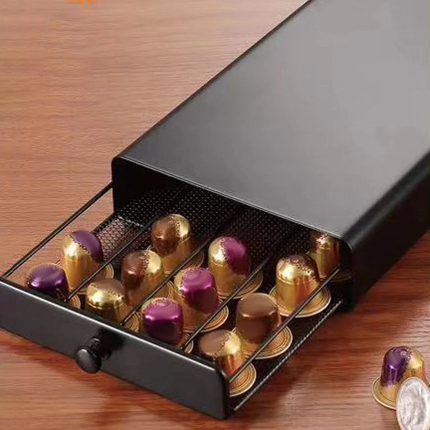 Een zwarte koffiecups organizer met een uittrekbare lade gevuld met goud- en paarse koffiecapsules, geplaatst op een houten ondergrond. Enkele losse koffiecapsules liggen naast de doos.